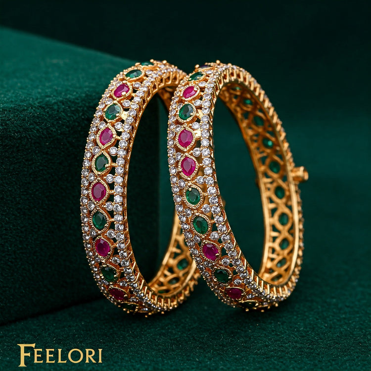 Pair of Feelori gold plated bangles intricately set with sparkling CZ, ruby-red, and emerald-green stones on green velvet - festive Indian jewelry.