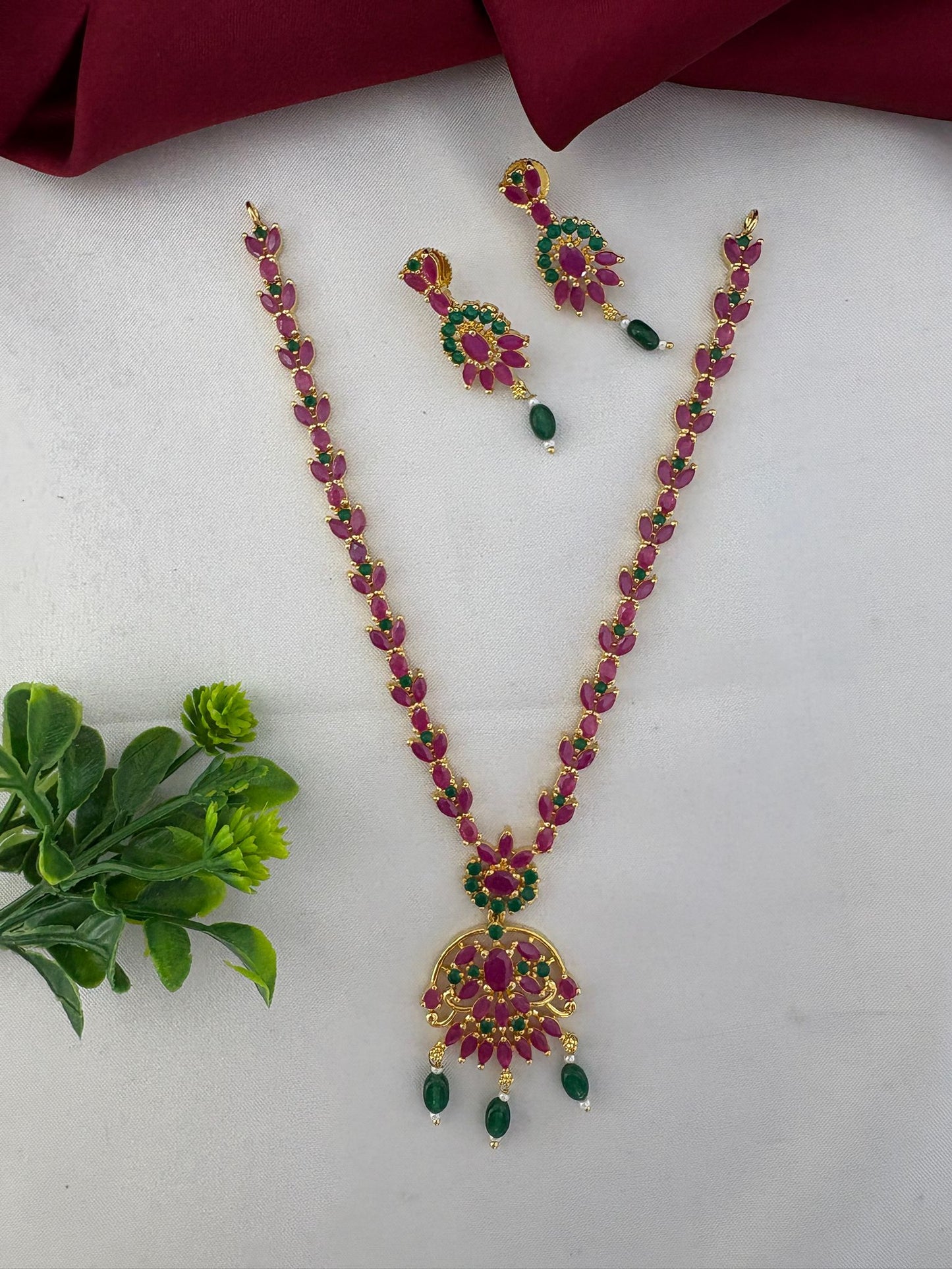 FeelOri Chettinad Bloom Ruby & Emerald Choker Set with matching earrings, elegantly styled with a green floral accent and maroon cloth.