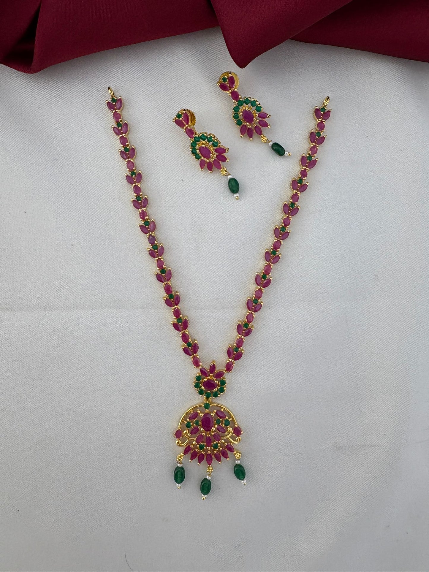 FeelOri Chettinad Bloom Ruby & Emerald Choker Set featuring necklace and earrings, top view on a white and rich maroon background.