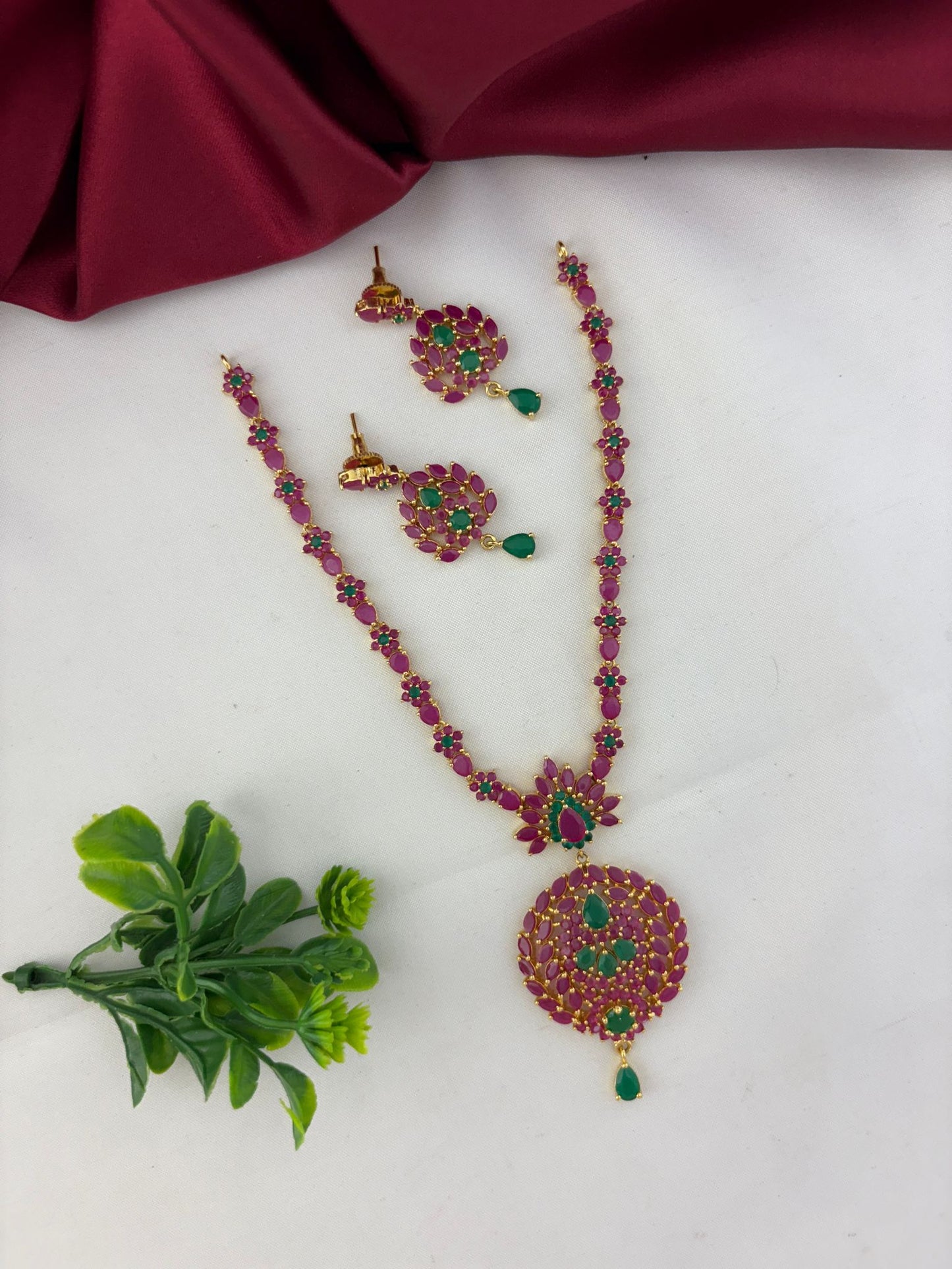 FeelOri Classic Duo Ruby & Emerald Short Necklace Set with earrings, styled with a touch of green next to the jewelry.