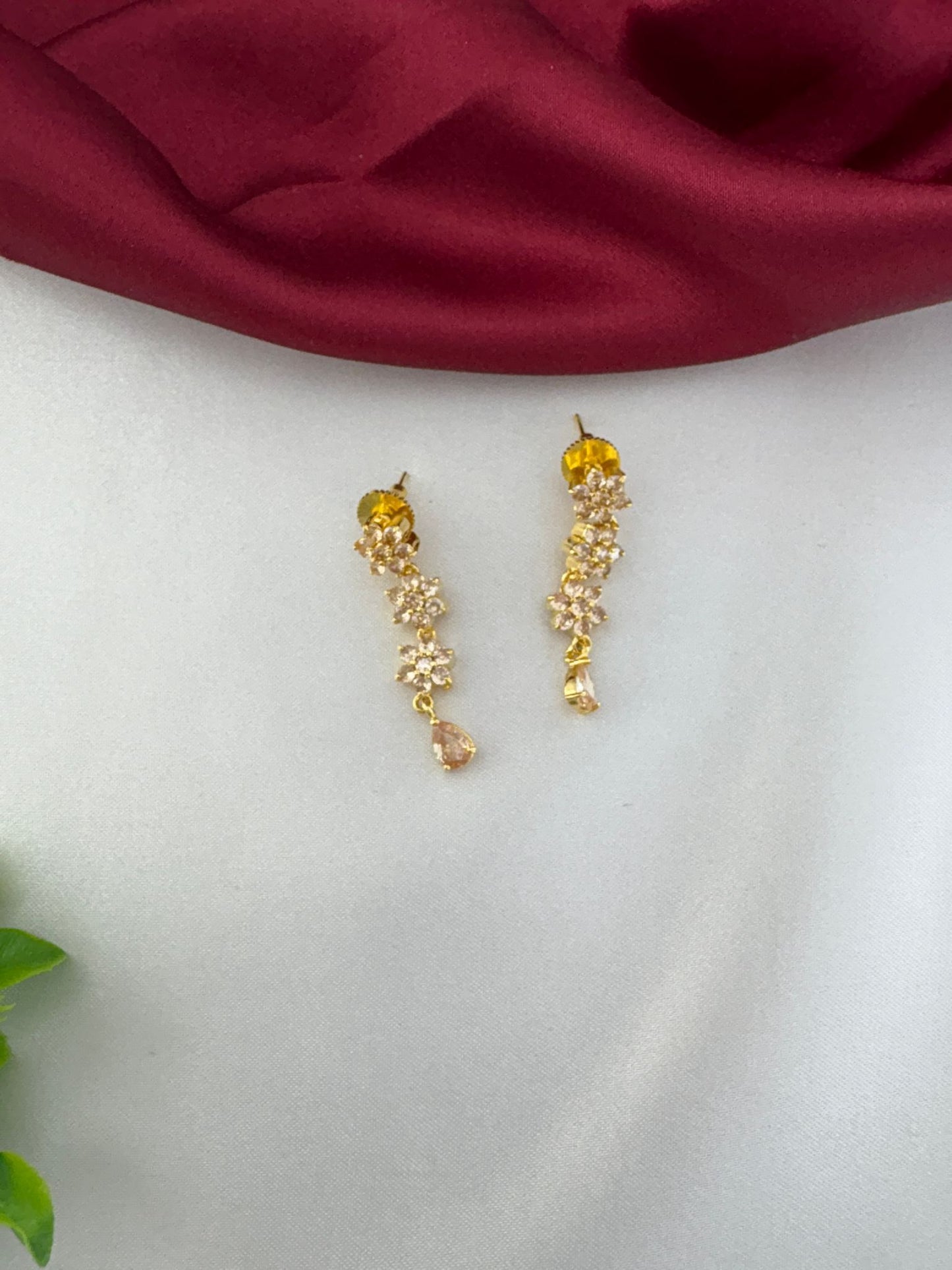 Detailed close-up of feelori Classic Gold Sparkle AD Earrings with floral design and a light champagne teardrop crystal, on white and burgundy background.