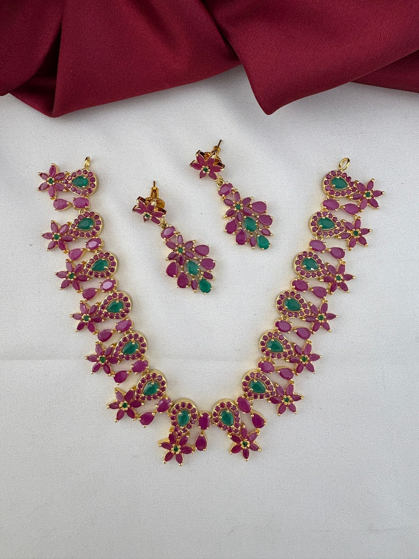 FeelOri Telangana Rani Ruby & Emerald Short Haar-Style Set - Full view of necklace and earrings on white and maroon background.