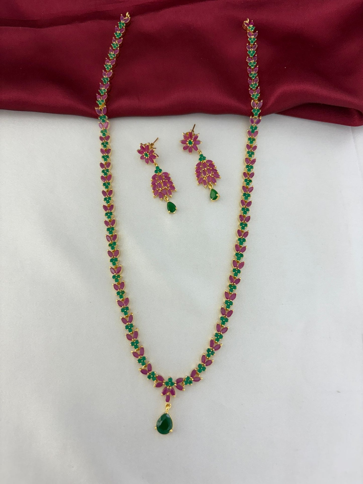 Feelorì Twin Souls Ruby & Emerald Alternating Long Necklace & Earring Set - Full view on maroon and white background.