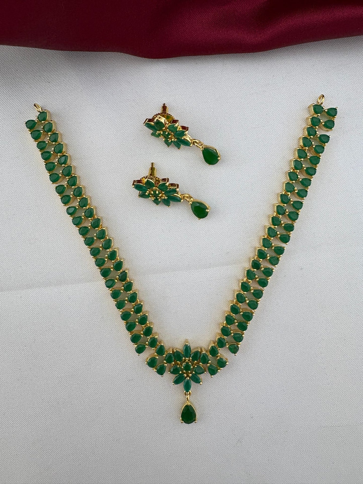 FeelOri Urban Oasis Minimalist Emerald Pendant Set featuring a necklace and matching earrings with vibrant green stones and gold-tone finish, displayed on a white and burgundy background.