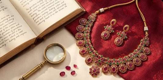 A flat lay photograph of a traditional gold-plated feelori ruby and emerald floral necklace set, similar to the FeelOri heritage collection, resting on antique brocade silk beside an open journal, a magnifying glass, and loose gemstones.
