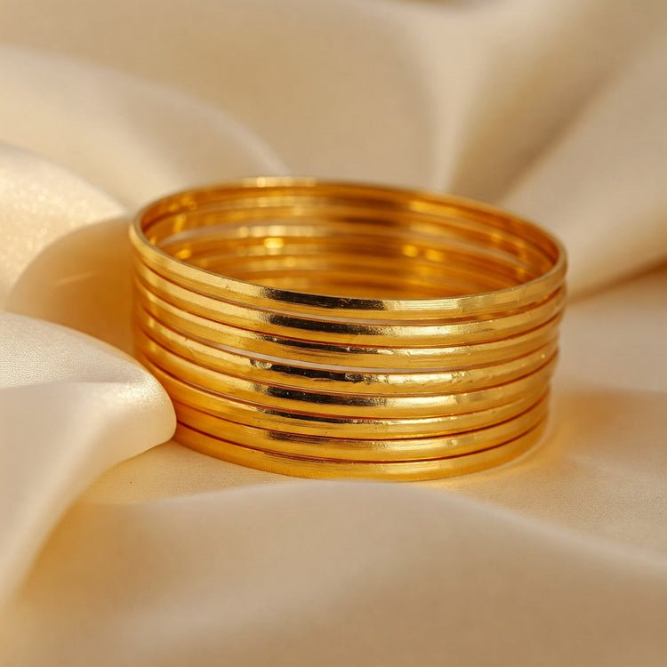 Stack of radiant Feelori gold plated bangles with a smooth finish, resting elegantly on soft ivory silk fabric - versatile Indian jewelry.