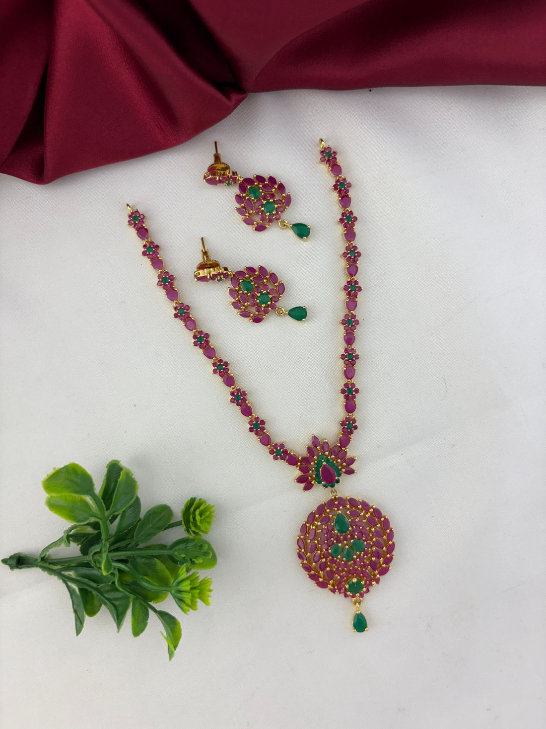 FeelOri Classic Duo Ruby & Emerald Short Necklace Set with earrings, styled with a touch of green next to the jewelry.