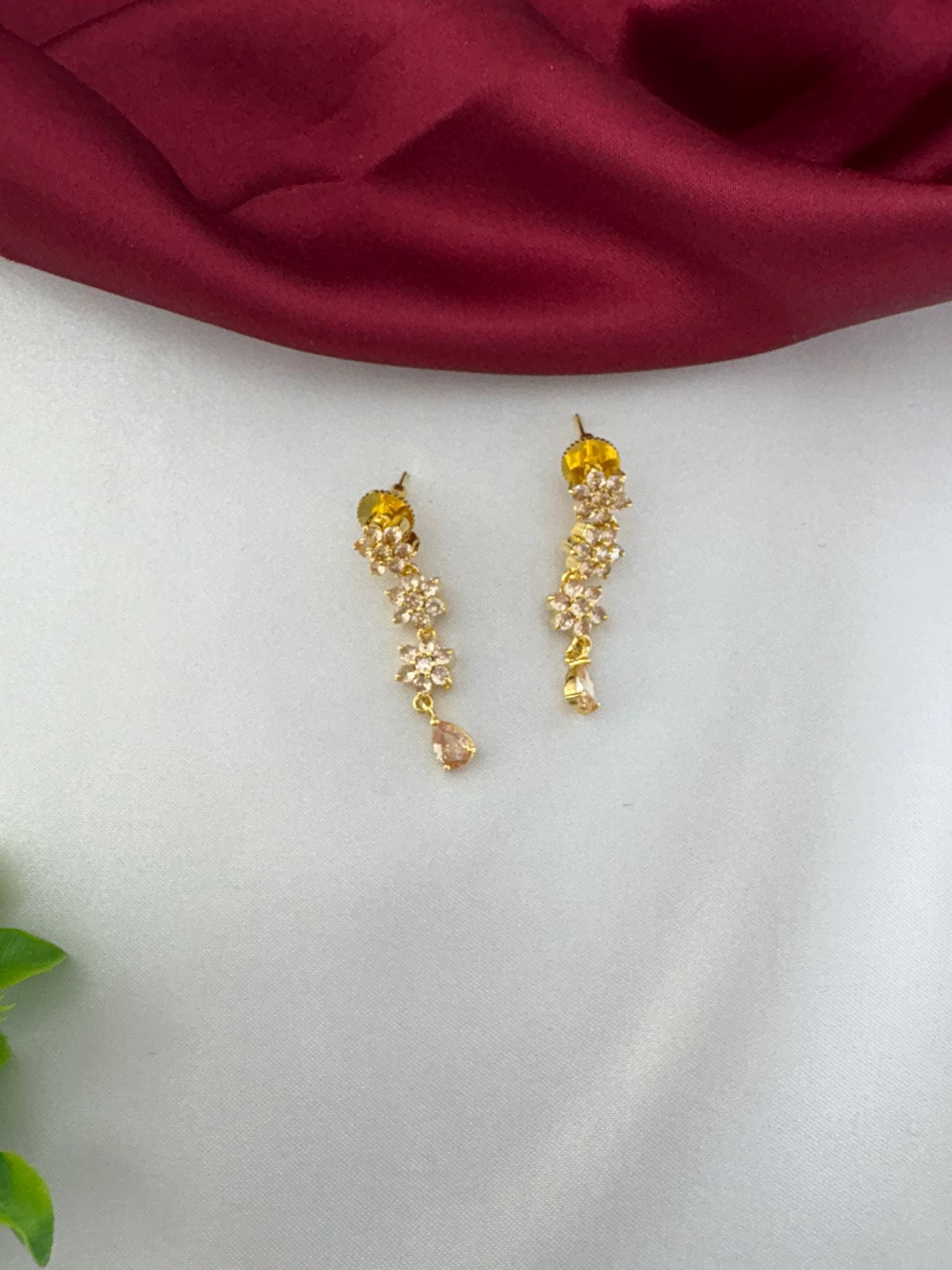 Detailed close-up of feelori Classic Gold Sparkle AD Earrings with floral design and a light champagne teardrop crystal, on white and burgundy background.