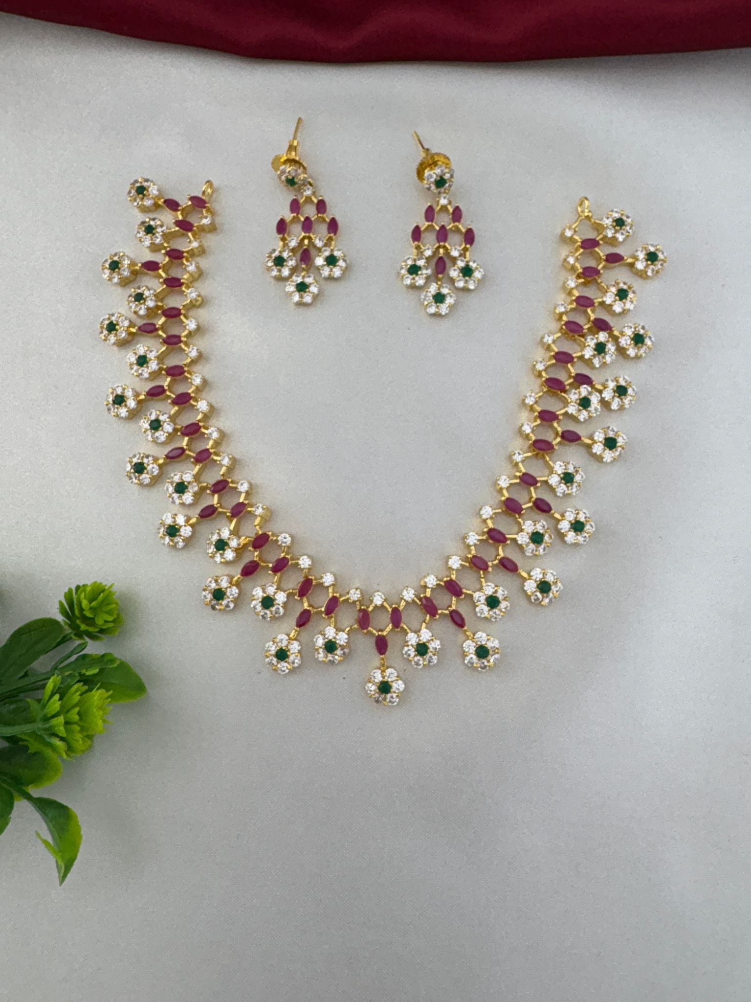 Styled view of FeelOri Jewel Tone Majesty Multi-Color AD Short Necklace Set with earrings, accented with a green plant.
