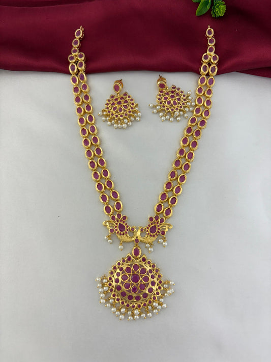 Top-down view of the FeelOri Royal Crimson Cascade Ruby Long Necklace and Earring Set. Highlights the straight alignment of the detailed ruby and pearl pendant and earrings. Set against white and maroon material.