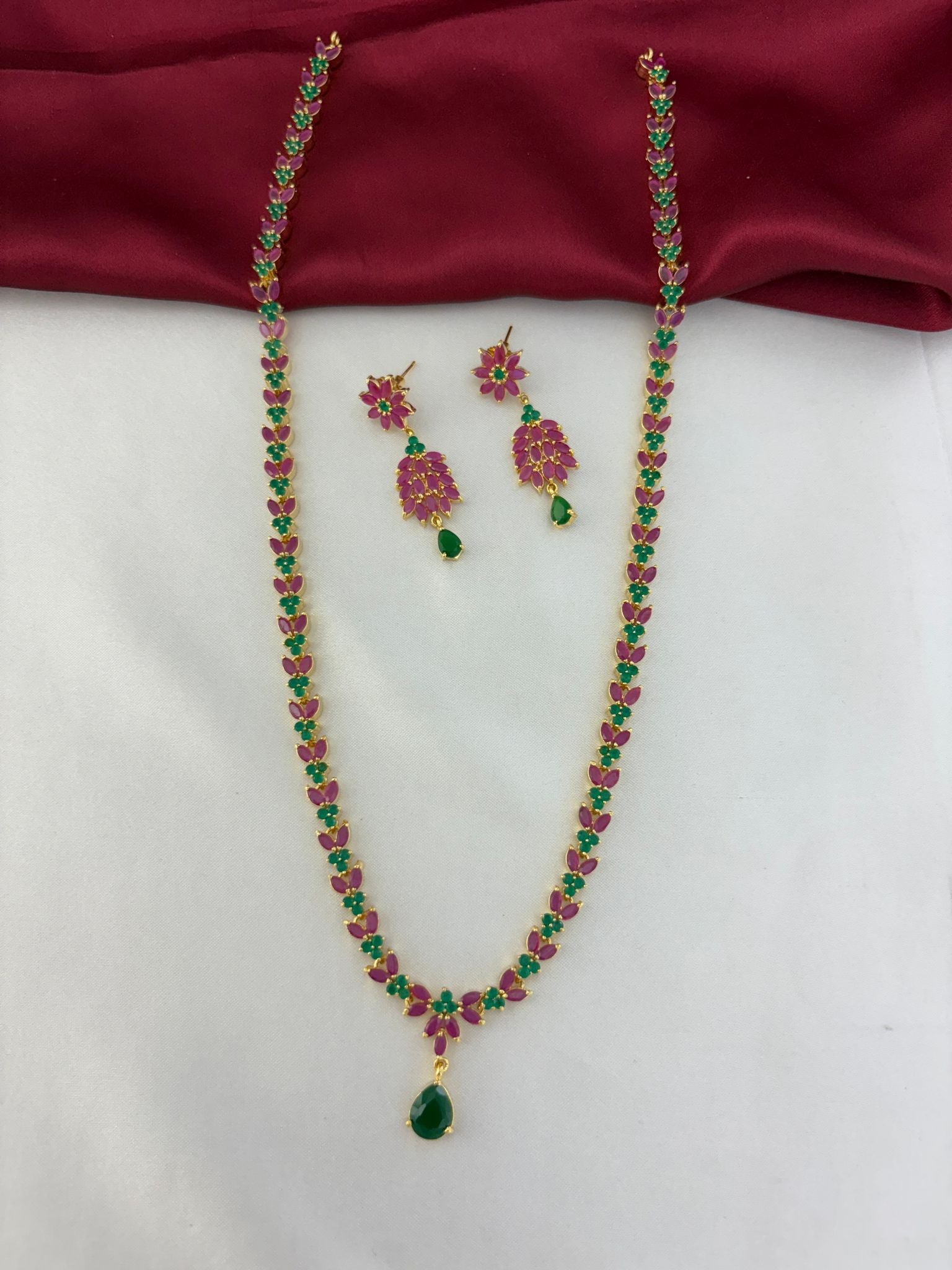 Feelorì Twin Souls Ruby & Emerald Alternating Long Necklace & Earring Set - Full view on maroon and white background.