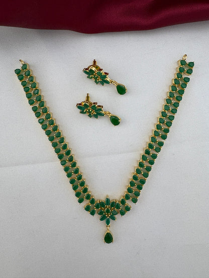 FeelOri Urban Oasis Minimalist Emerald Pendant Set featuring a necklace and matching earrings with vibrant green stones and gold-tone finish, displayed on a white and burgundy background.
