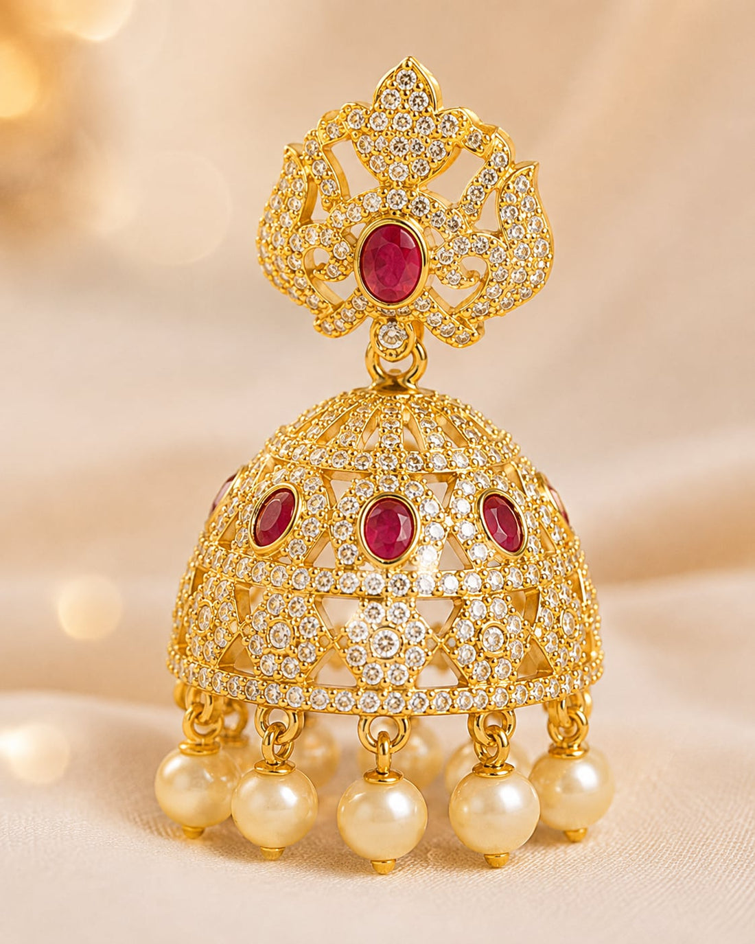Close-up of gold-plated ruby crown CZ jhumka earrings with pearl-look fringe showing crown-like top, ruby-red accents, clear CZ bell and pearl-look fringe